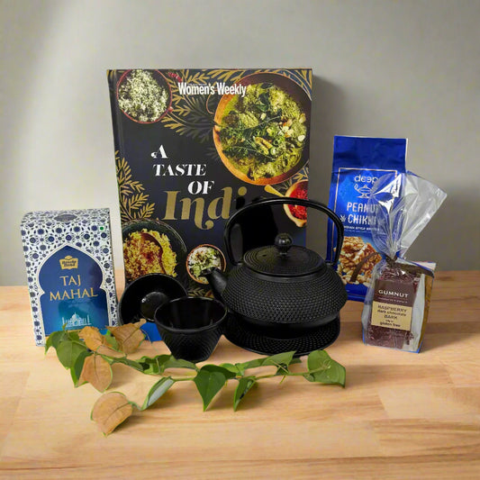 A gift set including a black hobnail cast iron teapot, a Women's Weekly cookbook, a camphor cheese board, assorted antipasto, and a box of heart-shaped chocolates, all displayed on a wooden surface with decorative leaves and a blue vase.